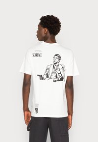 White cotton t-shirt featuring a black graphic of a seated man holding a gun, with "SCARFACE" text above and additional details below.