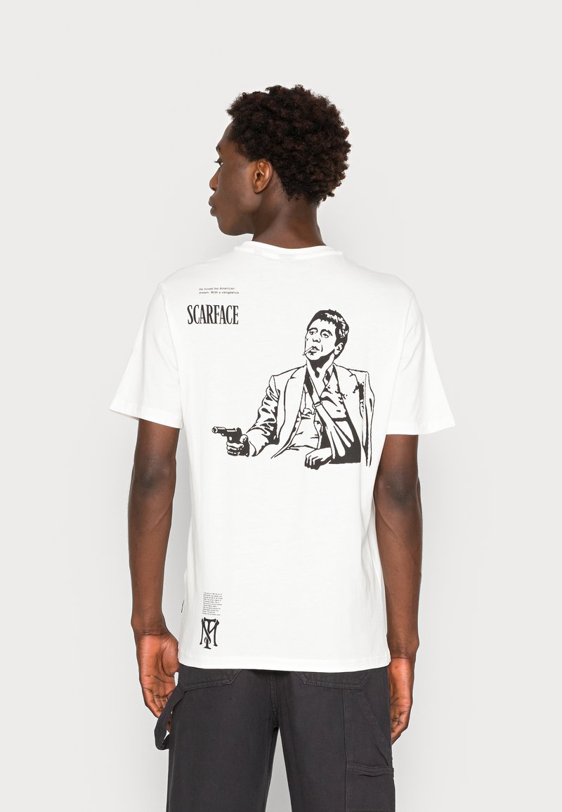 White cotton t-shirt featuring a black graphic of a seated man holding a gun, with "SCARFACE" text above and additional details below.