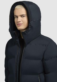Giacca piumino blu navy con cappuccio aderente, caratterizzata da una superficie liscia, chiusura frontale con zip e dettagli elastici attorno ai polsini e al cappuccio.