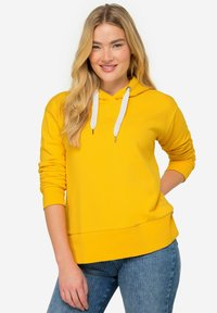 Felpa gialla con cappuccio regolabile, dettagli bianchi, polsini a costine e vestibilità rilassata. Abbinata a jeans blu con strisce verticali.