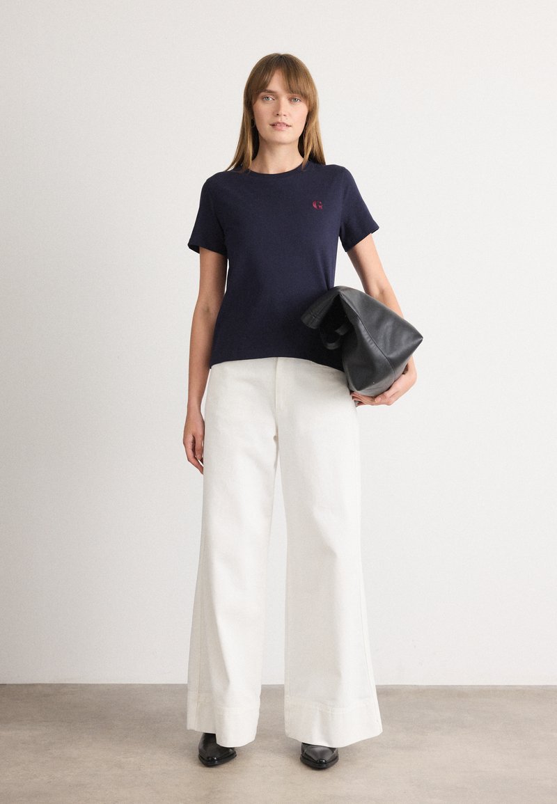 Marineblause mit kurzen Ärmeln und kleinem roten Logo, kombiniert mit hoch taillierten, weiten weißen Hosen. Model hält eine schwarze Schultertasche.