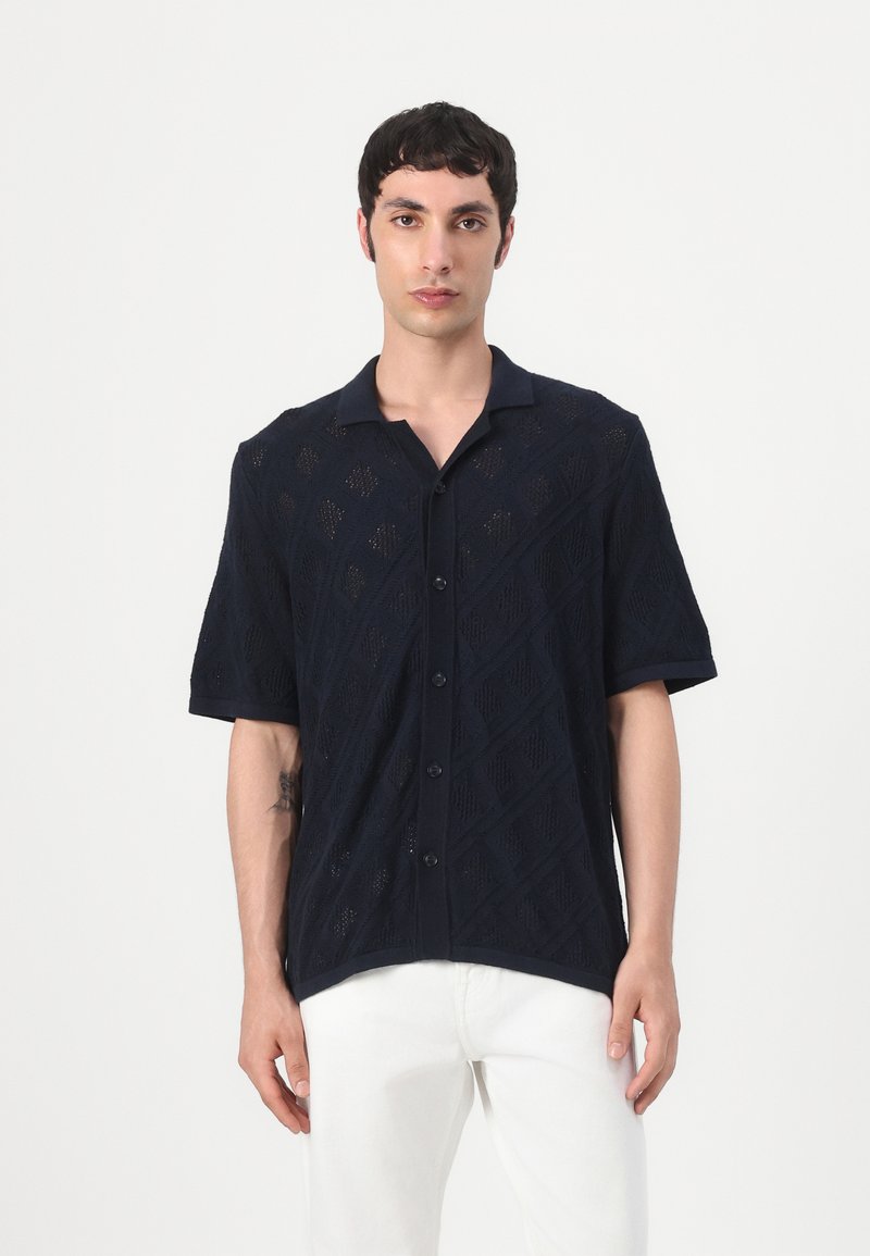 Cămașă navy cu mâneci scurte, din tricot cu model în diamond, cu nasturi în față și guler. Purtați cu pantalonii albi.
