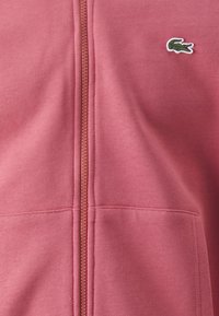 Felpa bordeaux con zip, realizzata in tessuto morbido, con logo coccodrillo verde ricamato sul lato sinistro e tasche a marsupio.