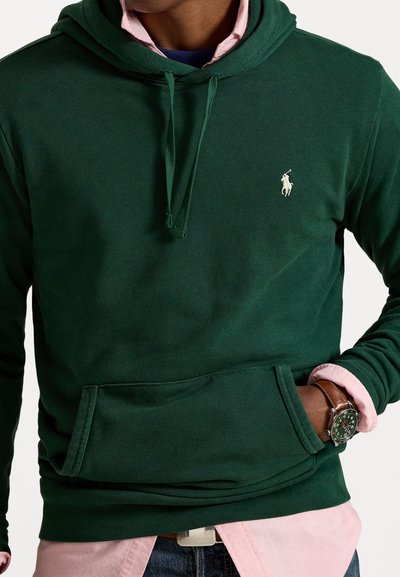 Donkergroene hoodie met een voorzak, trekkoorden en een klein wit logo. Draag over een lichtroze overhemd met kraag. Katoenmix stof.
