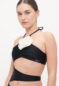 Čierny bikiny top s bielym textúrovaným kvetinovým akcentom, dizajn na krku, nariasená látka a logo na boku.