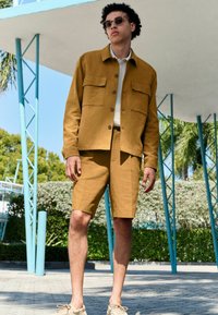 Sennepsfarvet bomuldsjakke med to forlommer, parret med matchende shorts. Modellen bærer solbriller og beige sneakers. Udemiljø.