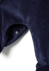 Indumento in velluto a coste blu navy con una superficie liscia, caratterizzato da chiusure a scatto e polsini a coste. Il tessuto presenta sottili righe verticali.