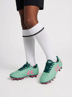 Person iført hvide knæstrømper og turkis fodboldstøvler med pink accenter, stående med den ene fod let hævet.