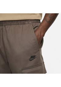 Ruka v kapse hnědých Nike tepláků s elastickým pasem a viditelným černým Nike logem na pravém stehně.