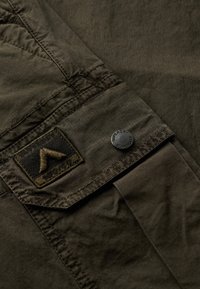 Olijfgroene shirt; lichtgewicht stof met een subtiel ruitpatroon; beschikt over een klepzak met een geborduurd logo en een ronde zwarte knop.