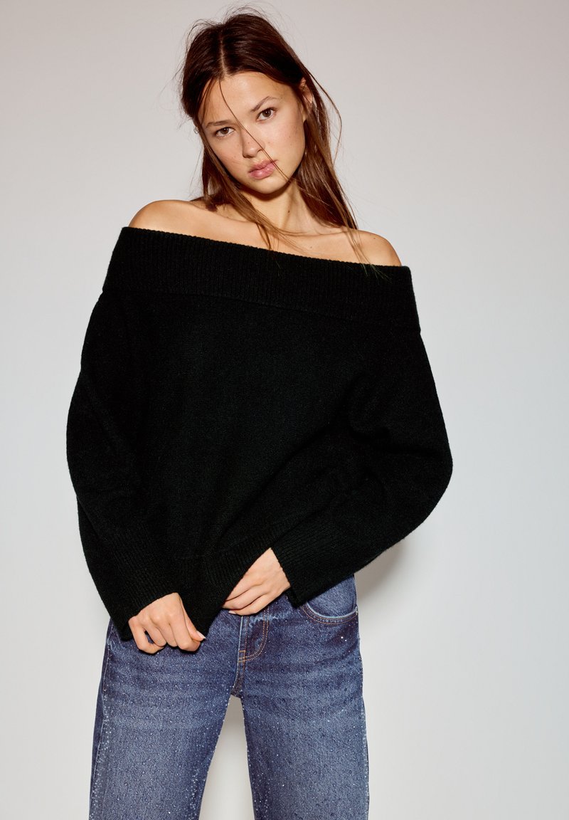 Zwarte off-shoulder gebreide trui met een brede geribbelde kraag, een losse pasvorm en lange mouwen, gecombineerd met lichtblauwe spijkerbroek.