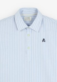 Camisa de rayas azul claro con cuello clásico, que presenta botones verdes y un pequeño detalle bordado de calavera y tibias cruzadas.