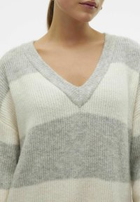 Pull léger en maille avec rayures crème et grises. Il présente un col en V et un tissu doux et texturé. Coupe décontractée avec épaules tombantes.