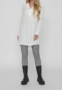 Vit skjorta med lång ärm och krage, nederkant längre bak. Kombinerad med grå rutiga byxor och svarta kraftiga stövlar.