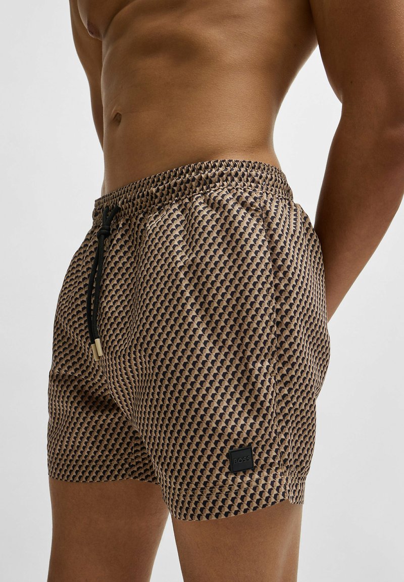 Men's swim shorts in patterned fabric, featuring a brown and black geometric design, elastic waistband, drawstring, and logo patch.