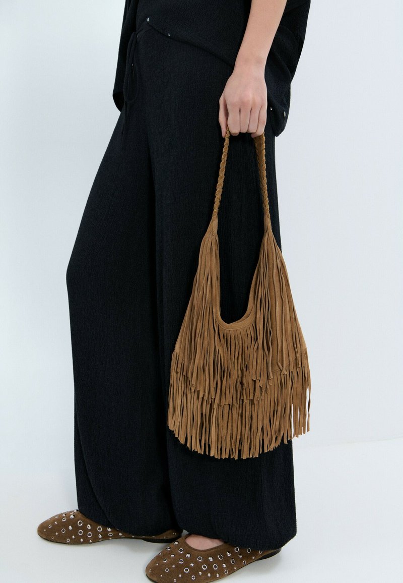 Mano sosteniendo un bolso de ante marrón con flecos, vistiendo pantalones negros de pierna ancha y zapatos de ante marrón con detalles de ojales plateados.