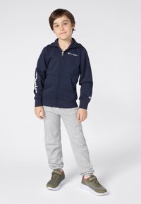 Mornarska zip-up dukserica s logotipom na prsima i rukavu, u kombinaciji s svijetlosivim trenirkama i tamnozelenim tenisicama. Glatka tkanina, opušteni kroj.