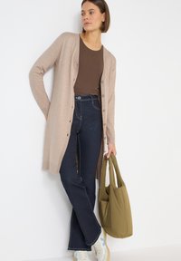 Světlý hnědý cardigan s knoflíky, nošený přes hnědý top, ve spojení s tmavými džíny s vysokým pasem a bílými teniskami, držící zelenou kabelku.