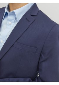 Granatowa marynarka o gładkiej fakturze, z pojedynczą kieszenią na piersi i subtelnymi przeszyciami. W zestawie z jasnoniebieską koszulą z kołnierzykiem.