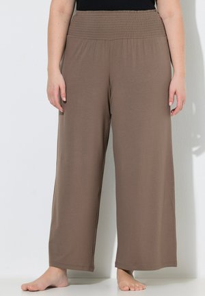 Persoon die losse, brede bruine broek draagt met een geplooide tailleband, blootsvoets staand tegen een eenvoudige lichte achtergrond.