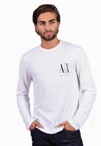 Vīrietis ar bārdu un uzspraustu matu griezumu, tērpies baltā, ar garajām piedurknēm, Armani Exchange kreklā ar melnu logo uz krūtīm, rokas tumšo džinsu kabatās.