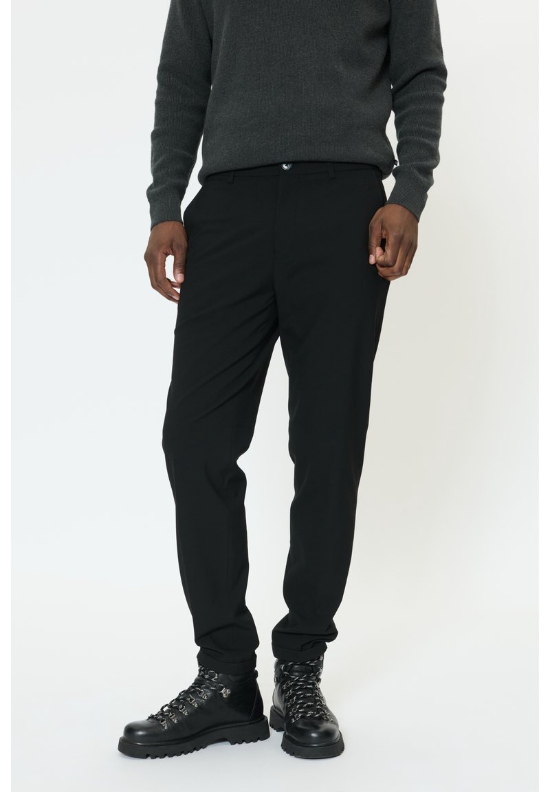Homme portant un pantalon noir fuselé, un pull à manches longues gris foncé et des bottes en cuir noires à lacets, debout sur un fond blanc.