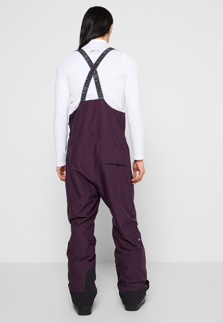 Fioletowe narciarskie ogrodniczki z regulowanymi czarnymi ramiączkami, dużymi kieszeniami, wzmocnionymi czarnymi mankietami oraz wytrzymałą fakturą materiału, idealne na zimowe aktywności.