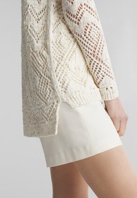 Sweter dzianinowy w kolorze kremowym z geometrycznym wzorem ażurowym, z długimi rękawami i bocznymi rozcięciami. Nosi się go na jasno kolorowej dopasowanej spódnicy.