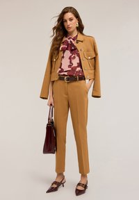 Krátká béžová bunda s kovovými knoflíky, blůza s květinovým vzorem, hnědý opasek, přiléhavé béžové kalhoty a vínová kabelka. Lodičky s špičatou špičkou.