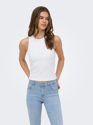 Kvinne med langt brunt hår iført en hvit ermeløs topp og lyseblå jeans, stående med hendene bak ryggen mot ensfarget bakgrunn.