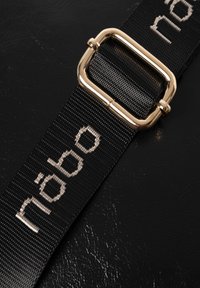 Cinturino intrecciato nero con il testo "näbo" in argento, fissato con una fibbia in metallo color oro su uno sfondo nero texturizzato.