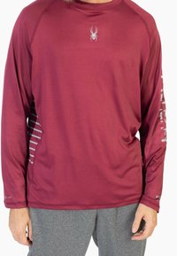 Bordowa sportowa koszulka z długim rękawem z szarym logo pająka na piersi i paskowanym wzorem po bokach. Gładka tekstura z przeszyciami na krawędziach.