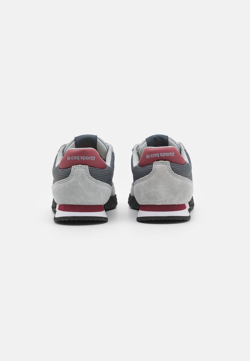 Baskets de sport grises avec une tige en suede et en mesh, dotées d'un accent rouge au talon et d'une semelle en caoutchouc noir. Étiquetées "le coq sportif" à l'arrière.