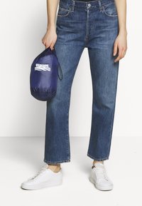 Jean en denim bleu, coupe droite, taille haute, avec des détails de décoloration subtils. Un sac compact navy avec un logo blanc est tenu à la main. Chaussures blanches.