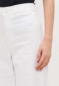 Pantalones de algodón blancos con una textura suave, que cuentan con cierre de botón, pasadores para cinturón y bolsillos laterales. Mano reposando sobre la tela.