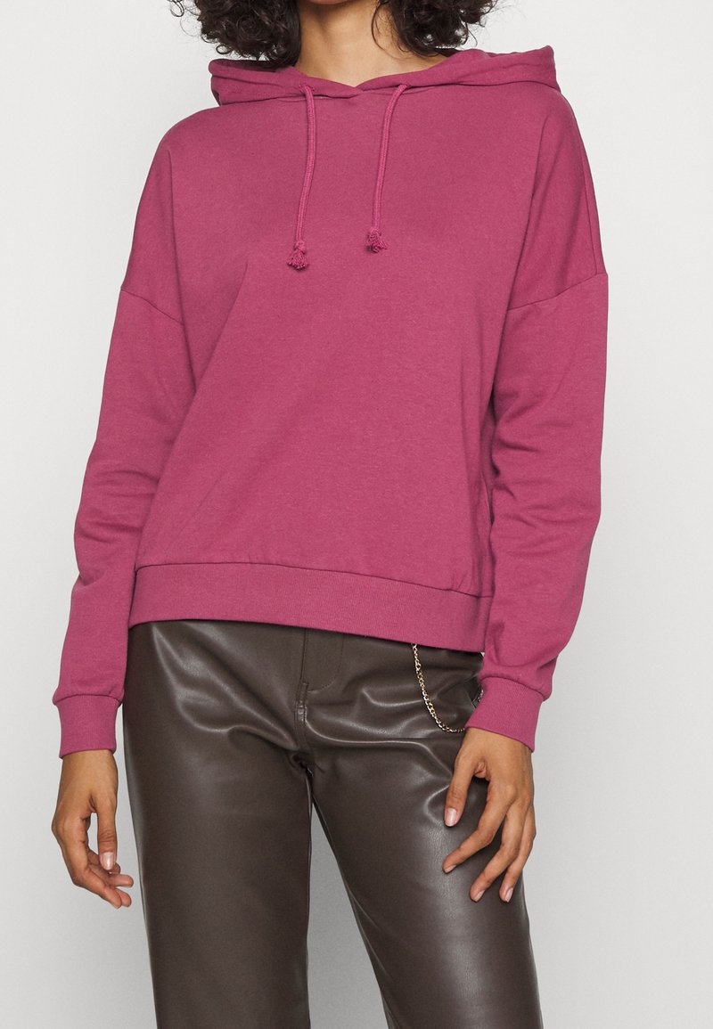 Felpa magenta con cappuccio regolabile, vestibilità rilassata e polsini e orlo a coste. Abbinata a pantaloni in eco-pelle marroni.