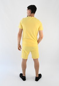 Camiseta amarilla y pantalones cortos a juego hechos de un tejido suave, con cuello de tripulación y un diseño minimalista. Llevado con zapatos negros.