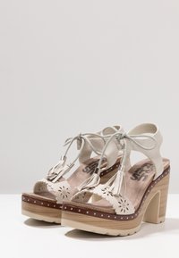 Högklackade sandaler i ljus beige mocka med dekorativa fransar, utskurna blommönster, träplattform och silverdetaljer.