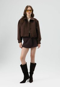 Bruine korte jas met grote kraag en knopen, gecombineerd met een donkerbruine minirok en zwarte suède laarzen tot aan de knie. Draagt een heldere gele zonnebril.