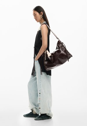 Young woman stands sideways wearing sleeveless black vest, wide light denim pants, black shoes, and carries large dark brown leather shoulder bag.