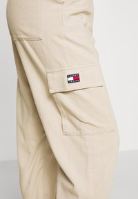 Beige cargo pants with a side flap pocket featuring a red, white, and blue Tommy Jeans logo patch.