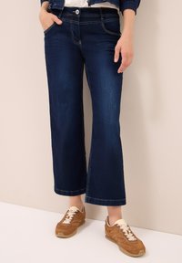 Persona con pantalones de mezclilla anchos de color azul oscuro y zapatillas marrones con cordones blancos, de pie contra una pared y un suelo beige.