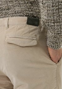 Béžové manšestrové kalhoty s náprsenkovou kapsou a zelenou koženou značkou na pásku. Viditelná žebrovaná textura.