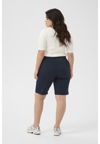 Navy blue hihnatut vaatteet suoraan leikkauksella ja takataskuilla, yhdistettynä kermaankeltaiseen lyhythihaiseen toppiin. Mallilla on päällään neutraalit lenkkurit.