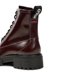 Botín de cuero burdeos con suela gruesa negra, tirador negro etiquetado "HUGO" y cordones negros visibles sobre un fondo blanco.