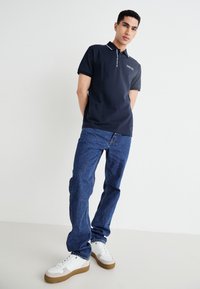 Jūras spēku polo krekls ar šauru piegriezumu, baltu apkakles akcentu un logotipa detaļu, kombinēts ar ziliem džinsiem un baltiem sporta apaviem ar brūnu zoli.