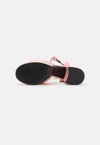 Sandale à talon haut rose vue de la semelle, avec un talon et l'avant-pied texturés noirs, et une bride à la cheville avec boucle, sur fond blanc.