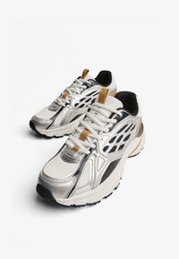 Para białych i srebrnych butów do biegania z czarnymi podeszwami i brązowymi uchwytami, ustawionych pod kątem obok siebie na białym tle.