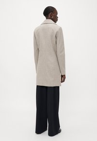 Beige wollen blend jas met een gestructureerd ontwerp, met een gesneden kraag en lange mouwen, getoond vanuit de achterkant. Gecombineerd met brede zwarte broeken.