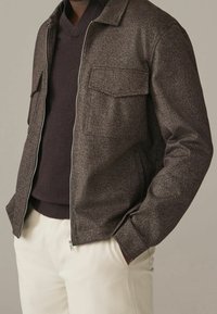 Bruine jas met visgraatmotief, twee borstzakken, ritssluiting aan de voorkant en opstaand kraag, gecombineerd met een donkere trui en lichte broek.